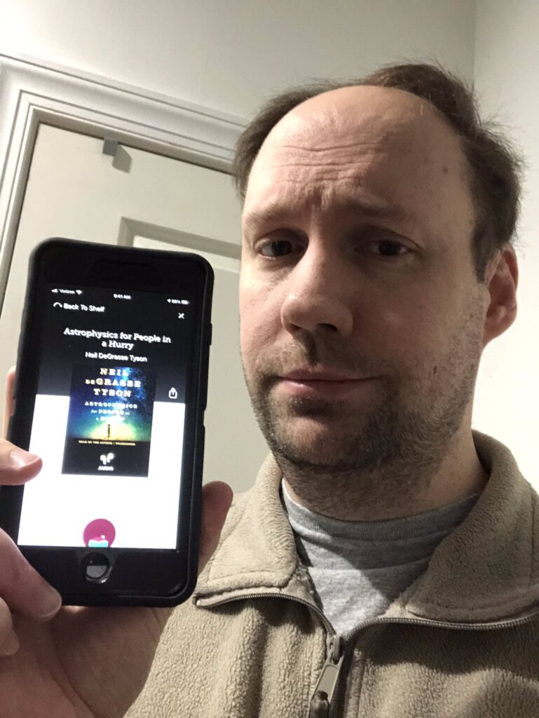 Matt holding up a phone displaying an audiobook of "Astrophysics for People in a Hurry"